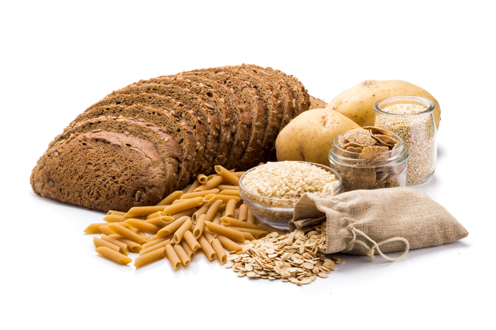 A variety of whole grains and starchy foods are displayed, including sliced brown bread, pasta, potatoes, oats, and jars of cereal and seeds, arranged on a white background.