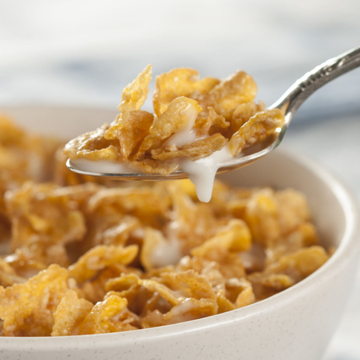 A spoonful of golden cornflakes topped with creamy milk is lifted from a bowl filled with more cornflakes, set against a softly blurred background.