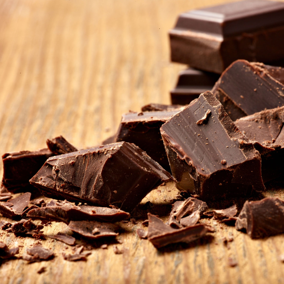 Chunks of dark chocolate are scattered on a wooden surface, with some pieces broken and others intact, showcasing their rich texture and deep color.