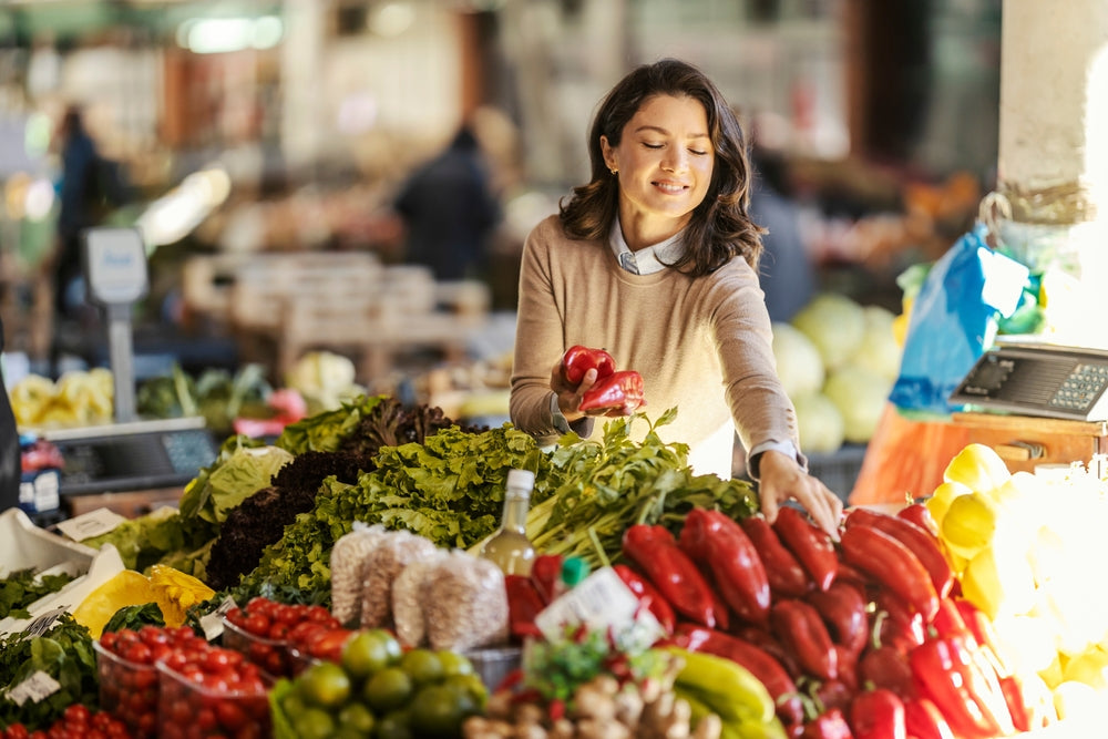 A woman in a beige sweater smiles while selecting red peppers from a vibrant display of fresh vegetables at a bustling market.