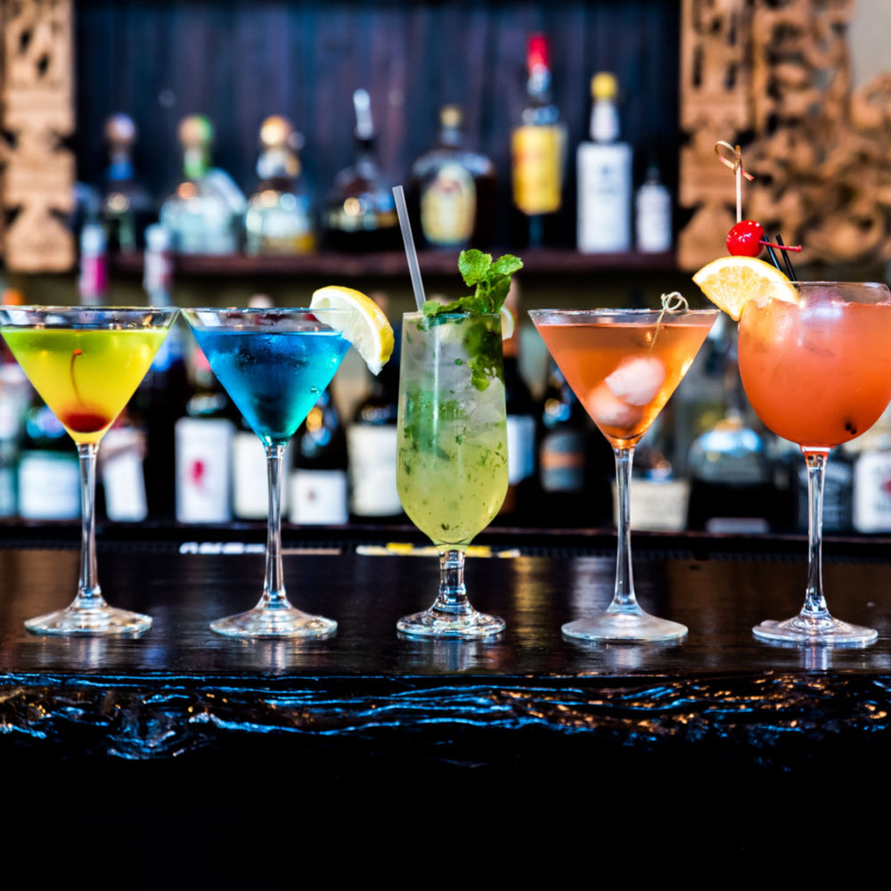 Five colorful cocktails are displayed on a bar counter, featuring a yellow drink with a cherry, a blue drink, a green mojito with mint, an orange drink, and a pink drink garnished with an orange slice and cherry, with bottles in the background.