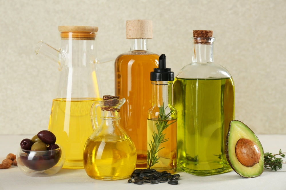 A collection of various glass bottles filled with different oils, including olive and avocado oil, is displayed alongside a bowl of olives and an avocado half on a light surface.