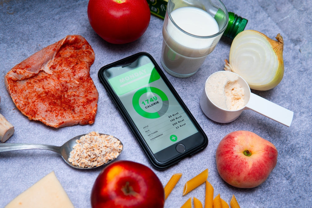 A smartphone displaying a calorie tracker sits among various food items, including a raw steak, apples, oats, milk, an onion, and pasta, on a textured surface.