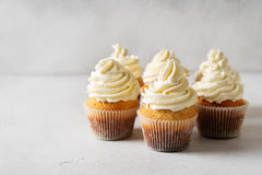 Five vanilla cupcakes with swirls of white frosting are arranged closely on a light gray surface, showcasing their golden-brown bases and decorative paper liners.