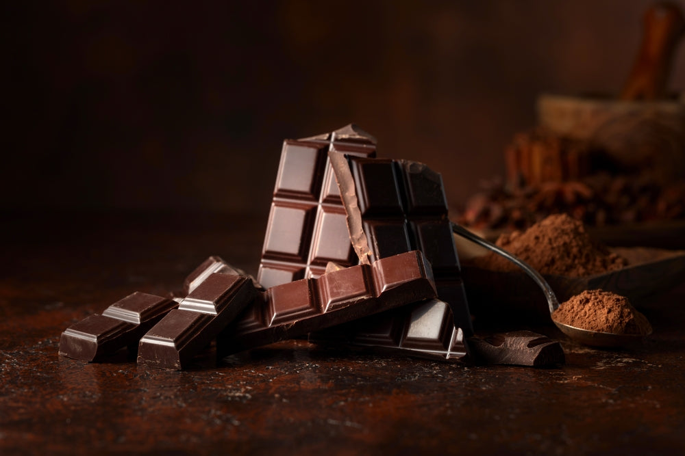 Dark chocolate bars are stacked on a rustic wooden surface, with cocoa powder and chocolate shavings scattered nearby, creating a rich, inviting atmosphere.