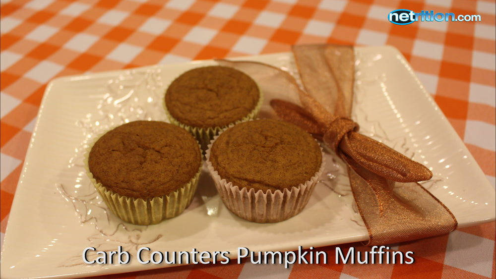 Three pumpkin muffins are arranged on a decorative white plate with a ribbon, set against a checkered orange tablecloth.
