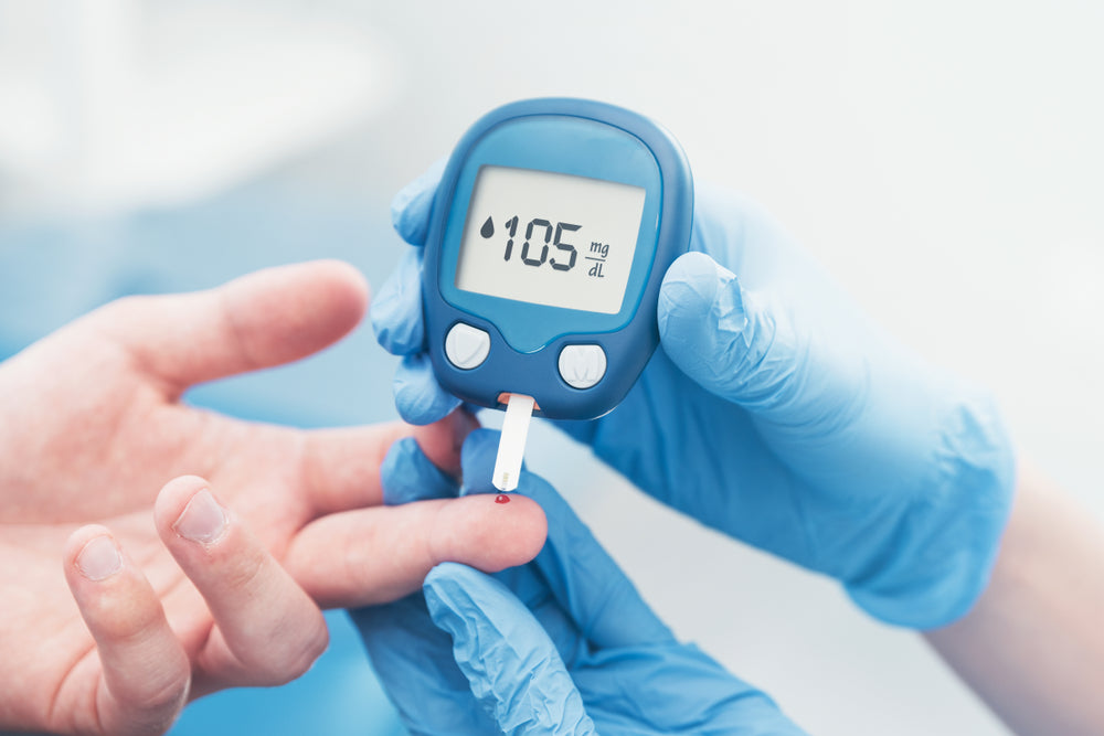 A healthcare professional in blue gloves holds a blood glucose meter displaying a reading of 105 mg/dL while preparing to test a patient's fingertip.