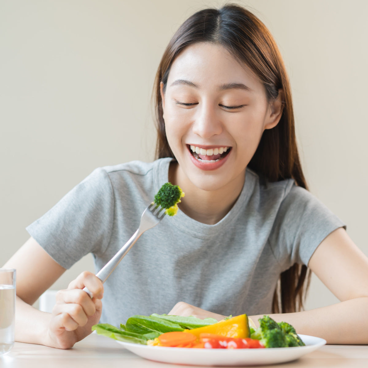 A young woman with long hair smiles joyfully while holding a fork with broccoli, sitting at a table with a colorful plate of vegetables and a glass of water beside her.