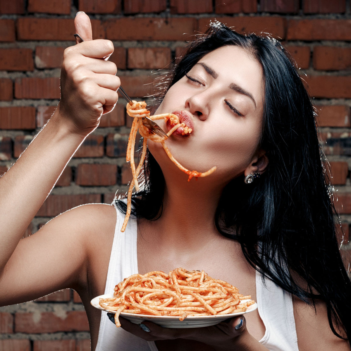 A woman with long black hair enjoys a plate of spaghetti, lifting a forkful to her mouth with her eyes closed, in front of a rustic brick wall.