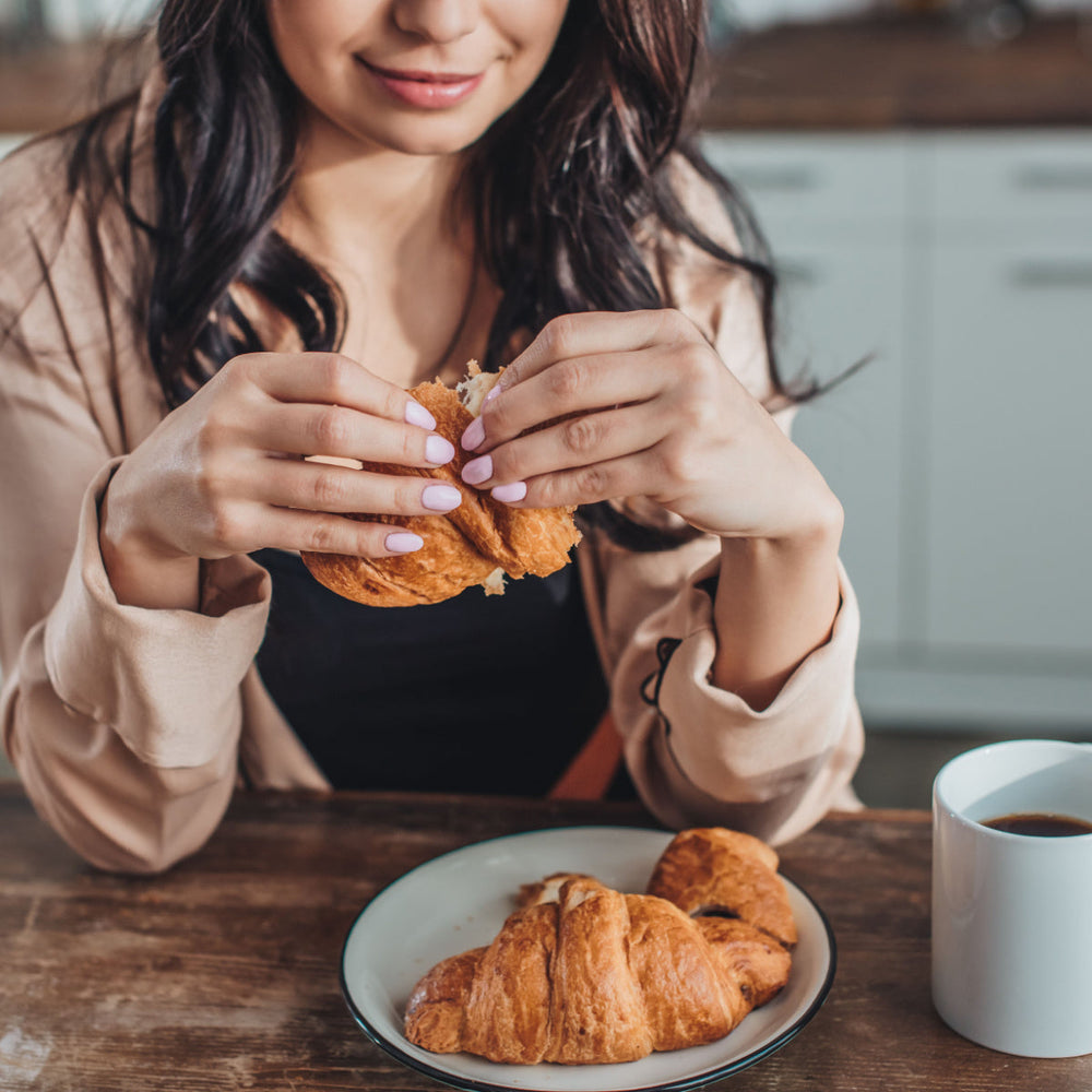 A woman with long dark hair smiles as she breaks a croissant in her hands, sitting at a wooden table with a plate of croissants and a cup of coffee in a cozy kitchen setting.