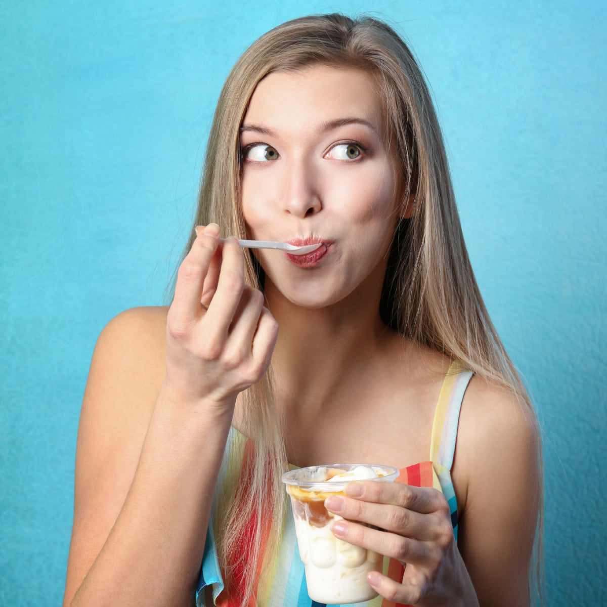 A young woman with long hair playfully eats from a plastic cup of dessert while smiling, set against a bright blue background.