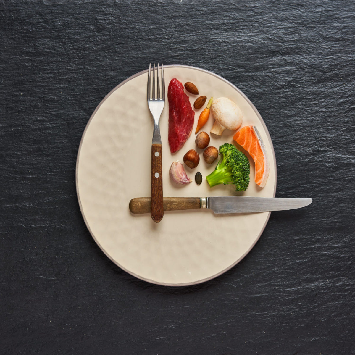 A beige plate with a fork and knife displays a variety of fresh foods, including pieces of meat, fish, vegetables, and nuts, arranged neatly against a dark slate background.