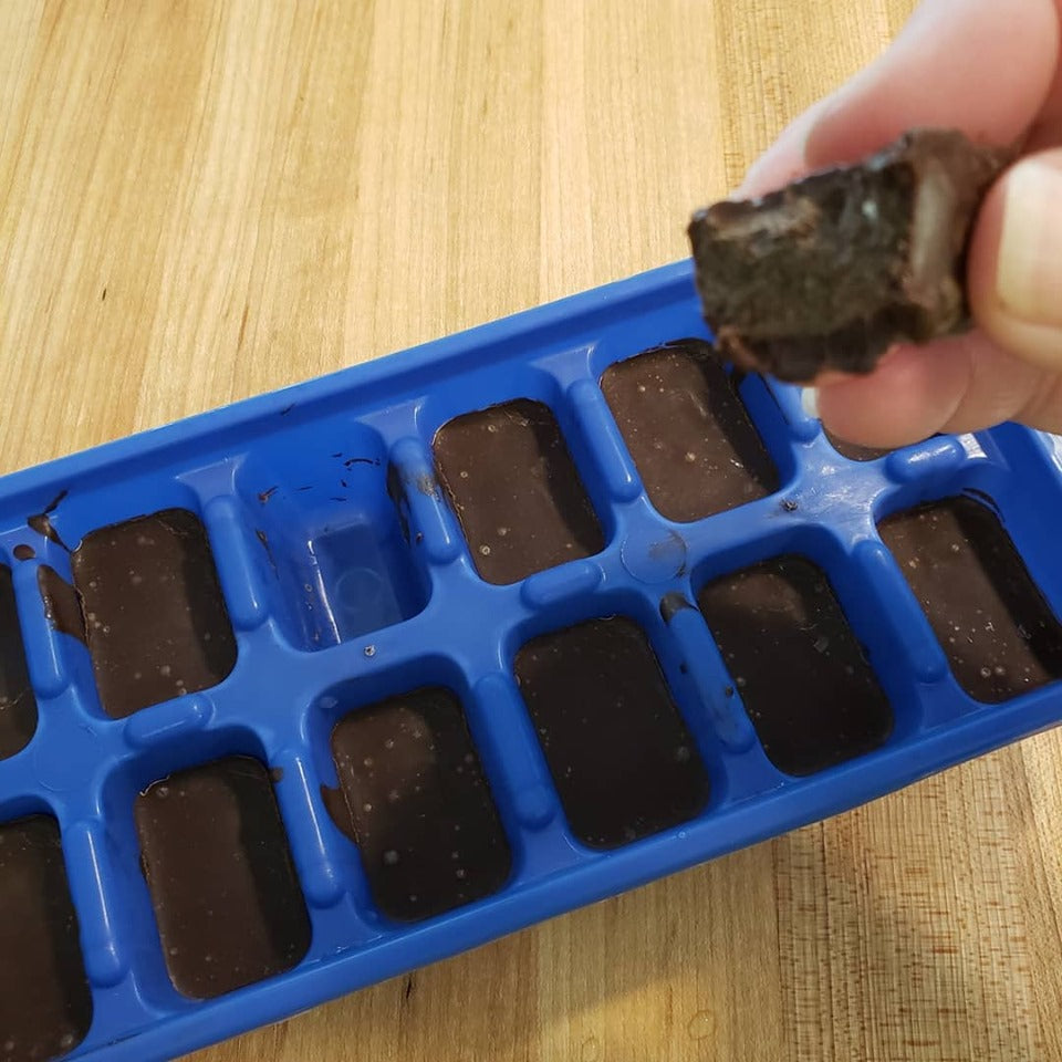 A hand holds a piece of chocolate above a blue silicone tray with several partially filled chocolate molds, resting on a wooden table.