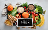 A variety of colorful fruits, vegetables, grains, and legumes are arranged on a table, with a chalkboard sign reading "FIBER" prominently displayed among them.