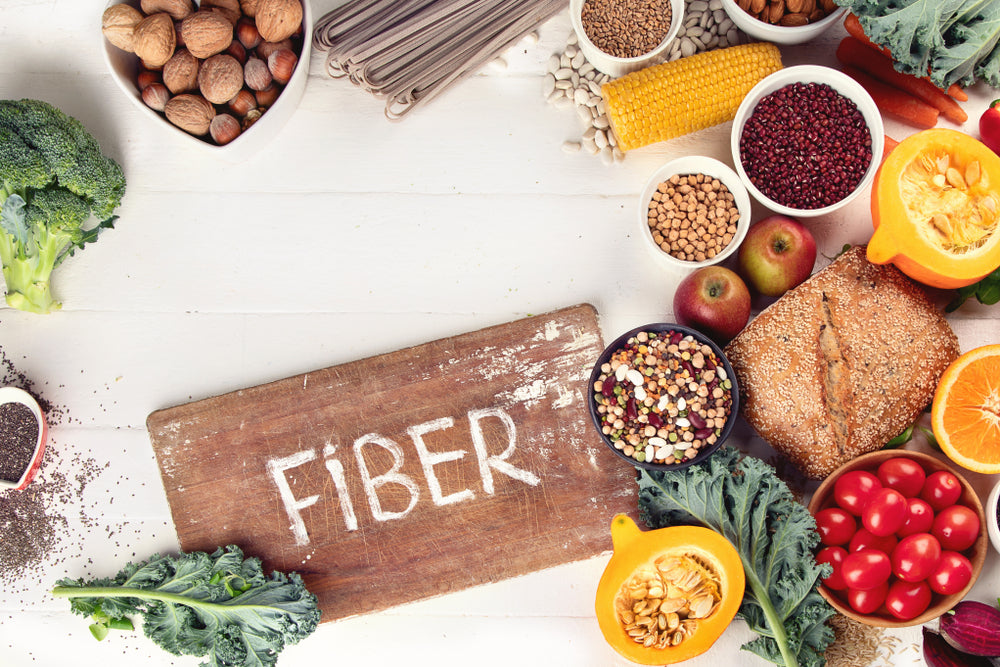 A wooden board labeled "FIBER" sits on a white table surrounded by various foods, including vegetables, fruits, nuts, grains, and legumes, highlighting healthy fiber-rich options.