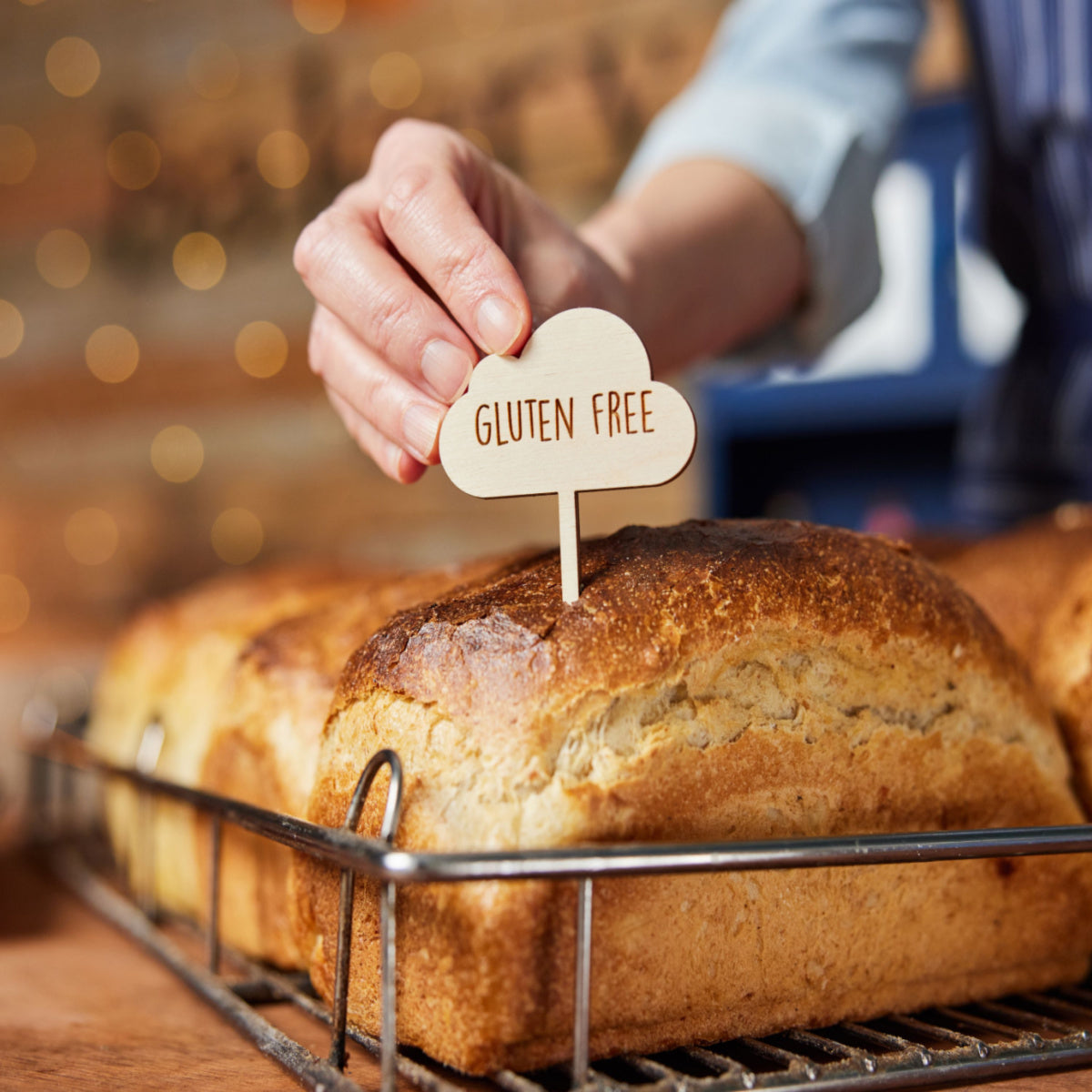 A hand places a wooden "Gluten Free" sign into a freshly baked loaf of bread, resting on a wire rack with a blurred, warm background of soft lights.
