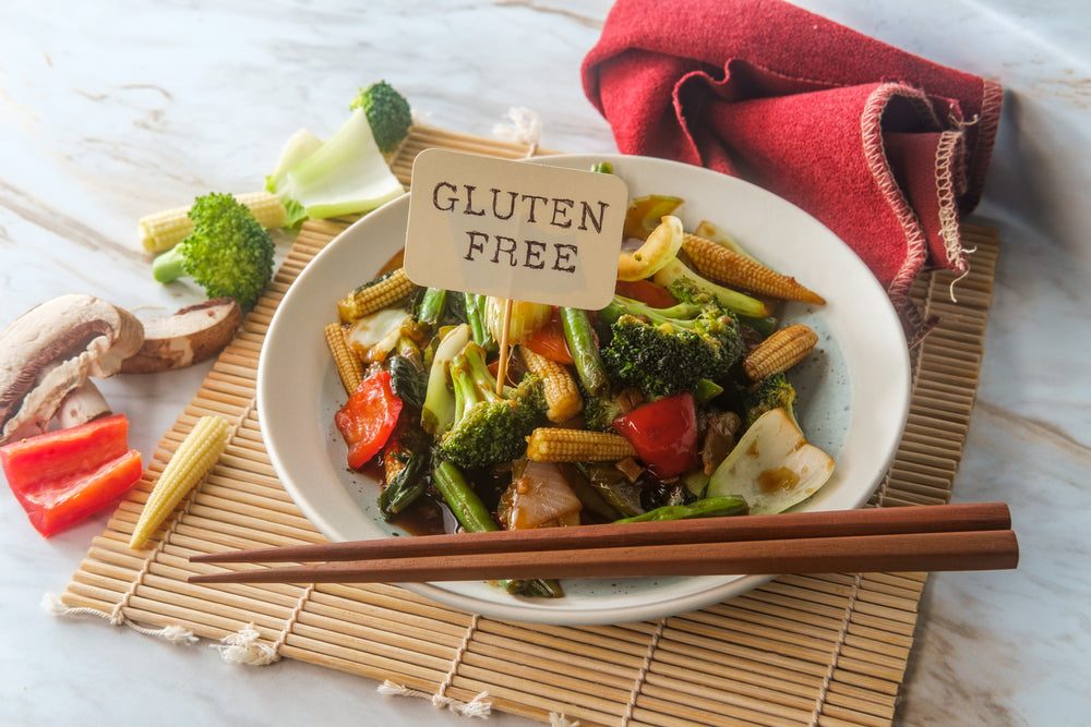 A bowl of colorful stir-fried vegetables, including broccoli and bell peppers, sits on a bamboo mat, with chopsticks resting beside it and a sign reading "GLUTEN FREE" displayed in the dish.