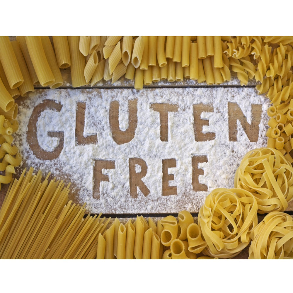 A wooden surface is covered with flour, displaying the words "GLUTEN FREE," surrounded by various types of uncooked pasta, including penne, fettuccine, and fusilli.