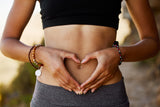 A person in a black crop top and gray leggings makes a heart shape with their hands over their bare stomach, standing outdoors with a blurred natural background.