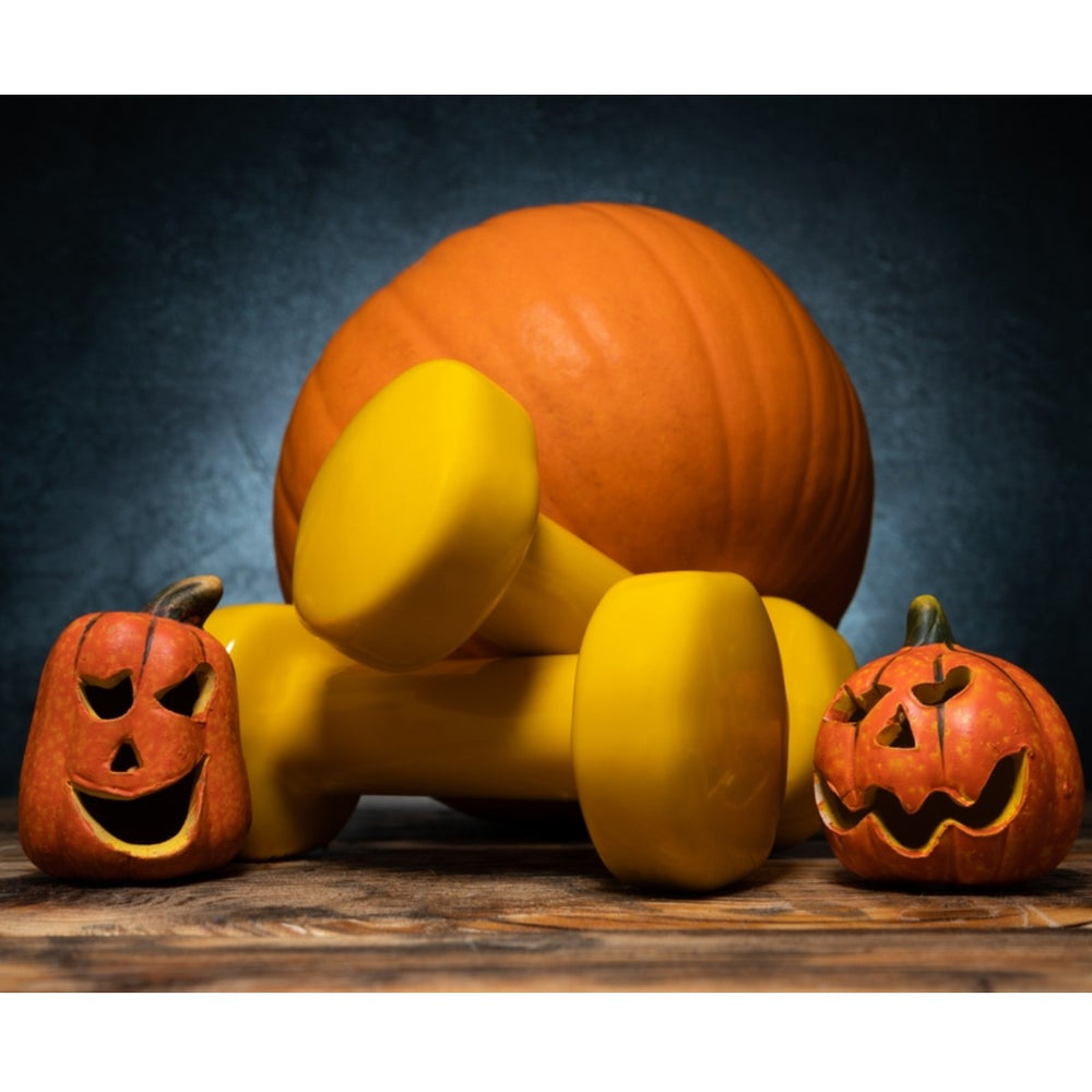 A large orange pumpkin rests behind two yellow dumbbells, with two carved jack-o'-lanterns displaying playful faces on a wooden surface against a dark background.