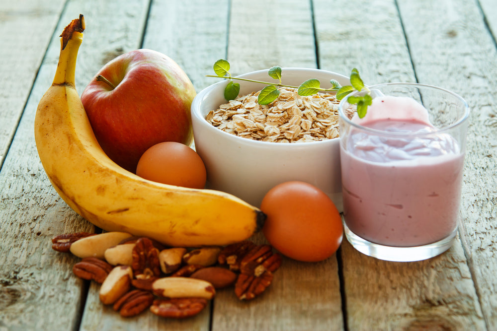 A banana, apple, two eggs, a bowl of oats topped with a sprig of green, and a glass of pink yogurt are arranged on a rustic wooden table, showcasing healthy breakfast options.