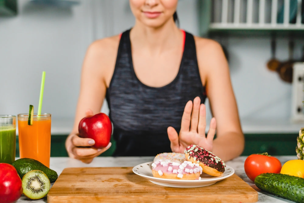 A woman in a tank top sits at a kitchen counter, holding an apple in one hand and gesturing with the other to refuse a plate of colorful donuts nearby, surrounded by fresh fruits and juices.