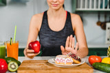 A woman in a tank top sits at a kitchen counter, holding an apple in one hand and gesturing with the other to refuse a plate of colorful donuts nearby, surrounded by fresh fruits and juices.