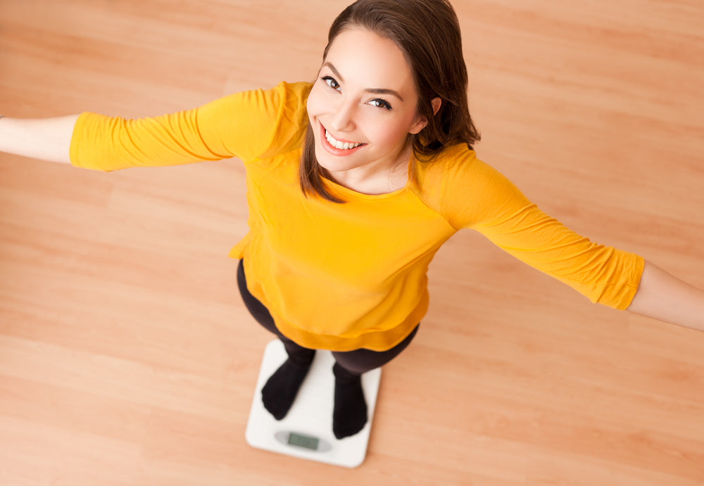 A woman in a yellow shirt stands on a bathroom scale with her arms outstretched, smiling happily, on a wooden floor.