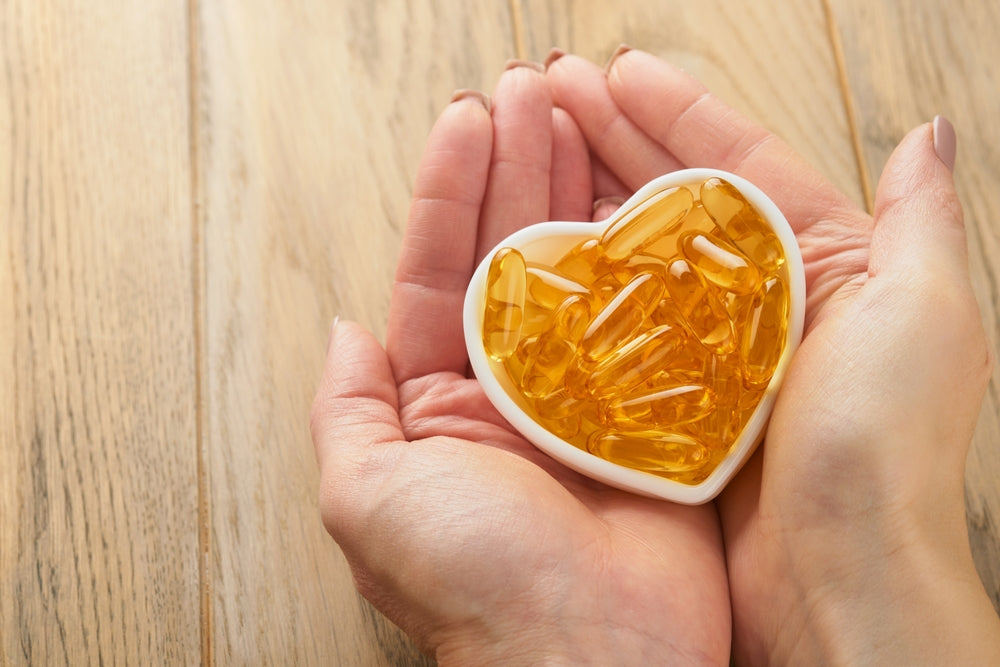 Two hands hold a white heart-shaped dish filled with golden gel capsules, resting on a wooden surface, symbolizing health and wellness.