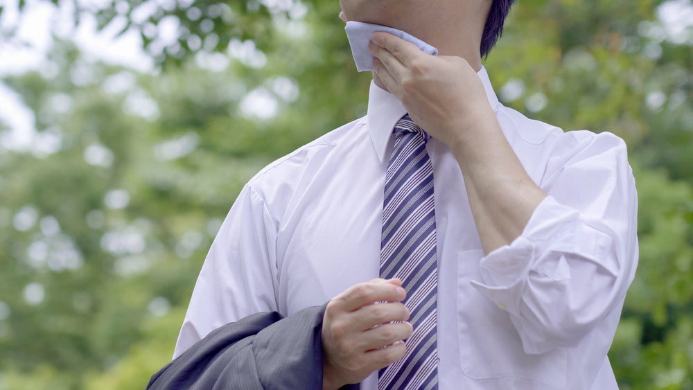 A man in a white dress shirt and striped tie wipes his neck with a cloth while standing outdoors in a green, leafy setting, appearing to cool off or freshen up.
