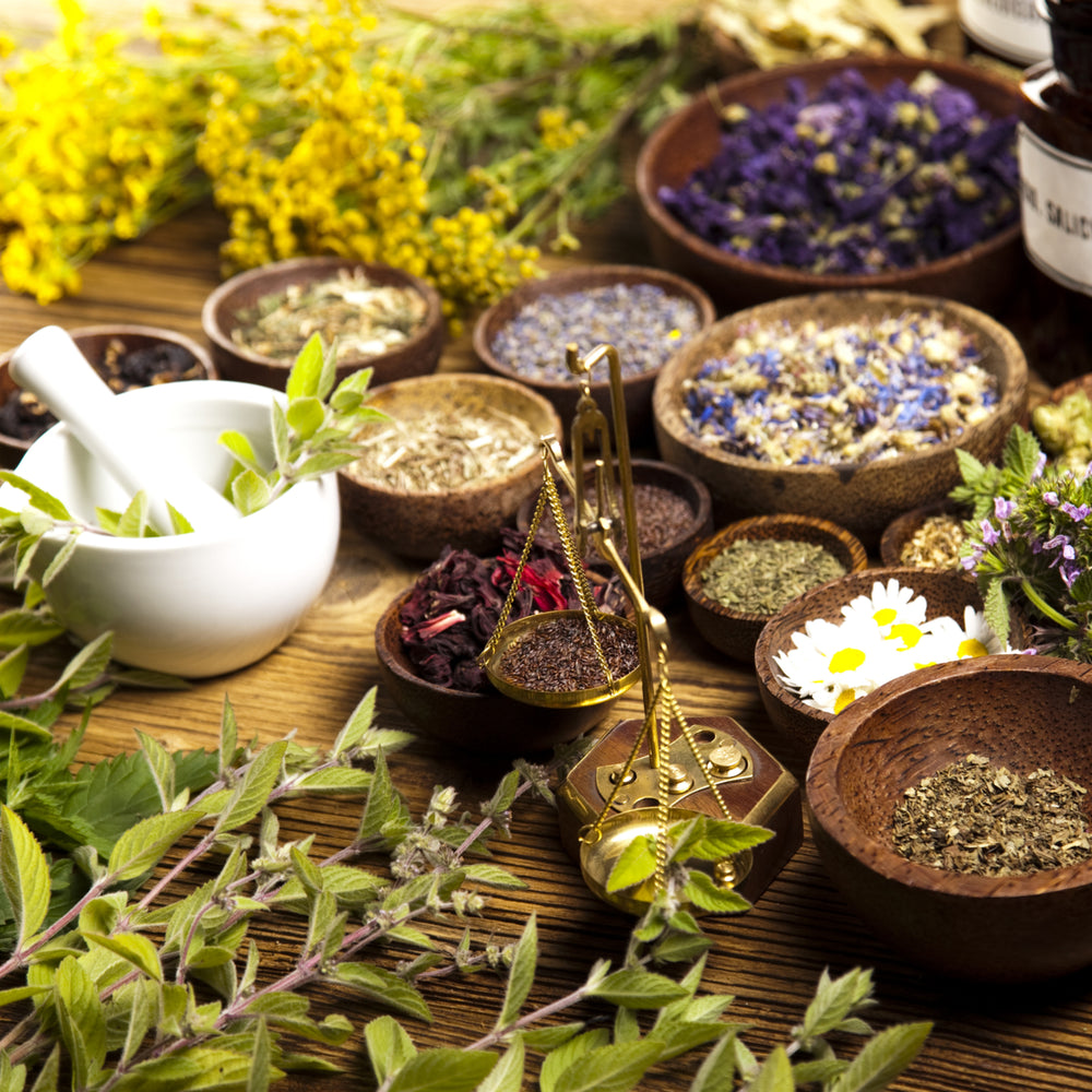 A wooden table is filled with various bowls of colorful herbs and flowers, a white mortar and pestle, and a small balance scale, creating a vibrant setting for herbal preparation.