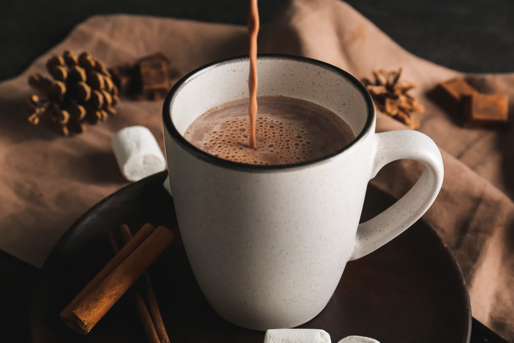 A white mug filled with hot chocolate is being poured, surrounded by pinecones, cinnamon sticks, marshmallows, and chocolate pieces on a brown cloth and dark plate.