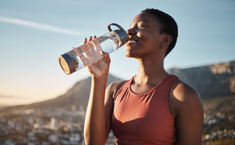 A woman with short hair smiles while drinking water from a clear bottle, set against a backdrop of mountains and a city during sunset.