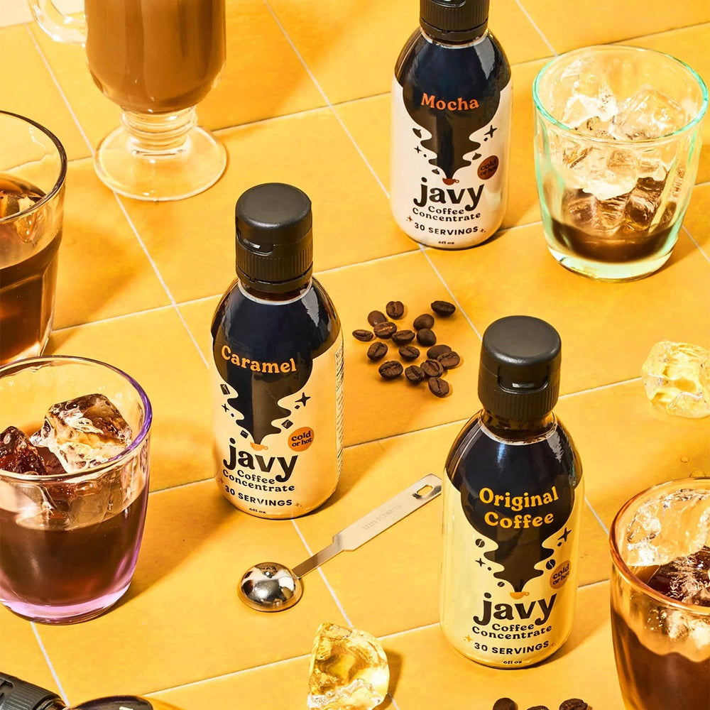 Three bottles of Javy Coffee concentrate—Caramel, Mocha, and Original—are displayed on a yellow surface surrounded by ice cubes, coffee beans, and glasses of iced coffee.
