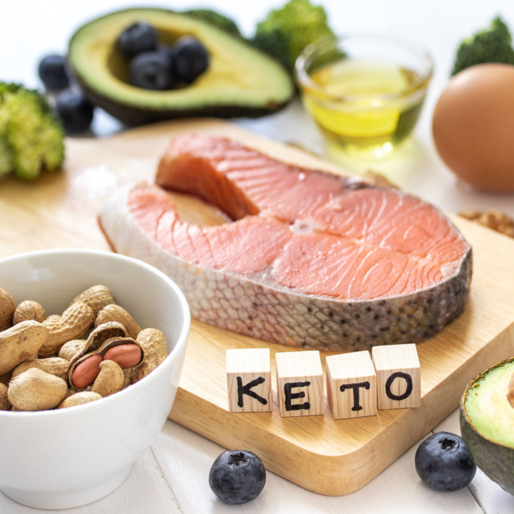 A wooden cutting board displays a salmon fillet, a bowl of mixed nuts, an avocado, blueberries, an egg, and olive oil, with wooden blocks spelling "KETO" in the foreground.
