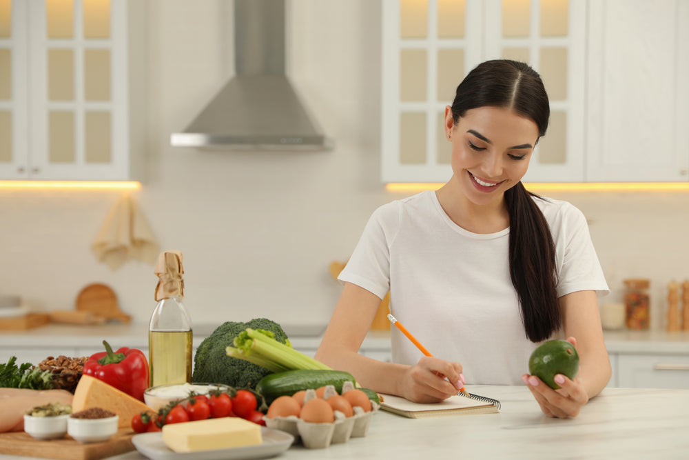 A woman in a white shirt smiles while writing in a notebook, surrounded by fresh vegetables, eggs, and cooking oil on a kitchen counter, suggesting meal planning for a ketogenic diet.