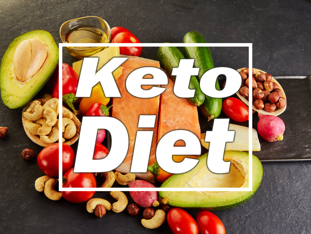 A variety of fresh foods including avocado, salmon, nuts, and vegetables are arranged on a dark surface, with the words "Keto Diet" prominently displayed in the center.