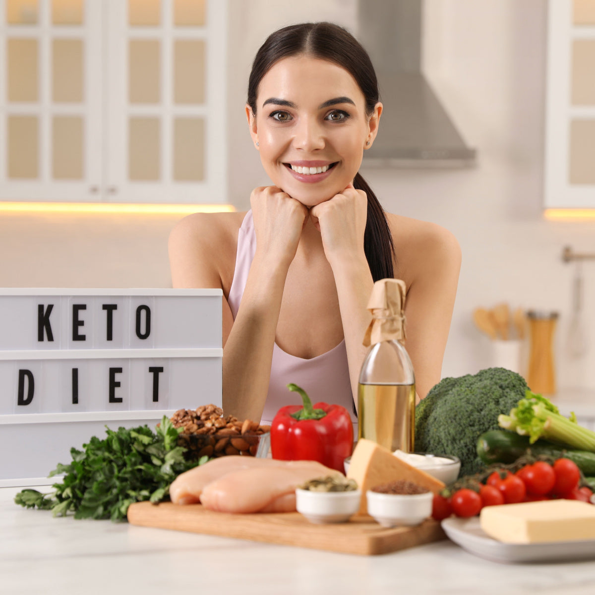 A smiling woman rests her chin on her hands, surrounded by fresh vegetables, meats, and oils, with a sign that says "KETO DIET" in a bright kitchen setting.