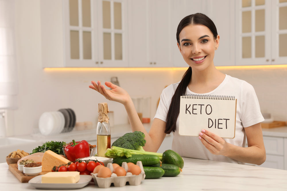 A smiling woman in a kitchen holds a notebook displaying "KETO DIET" while gesturing towards a variety of fresh vegetables, cheese, and eggs arranged on the counter.