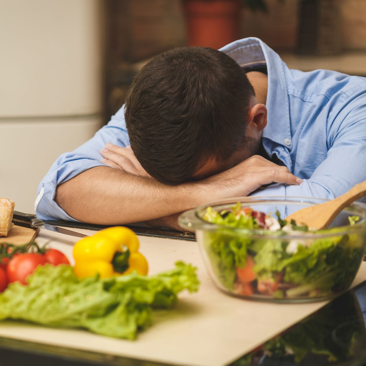 A man in a blue shirt rests his head on his arms at a kitchen counter, surrounded by fresh vegetables and a bowl of salad, appearing overwhelmed or frustrated.