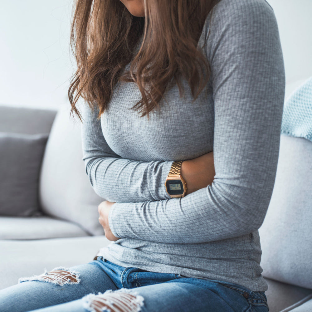 A woman in a gray long-sleeve shirt sits on a couch, holding her stomach with a pained expression, wearing a gold watch, with a cozy living room setting in the background.
