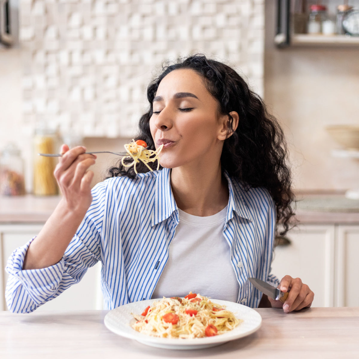 A woman with curly hair enjoys a forkful of spaghetti with cherry tomatoes, smiling with her eyes closed, seated at a kitchen table with a plate of pasta in front of her.