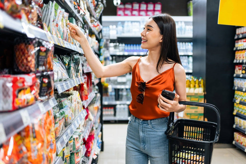 A woman in a red top smiles while reaching for a product on a grocery store shelf, holding a shopping basket in one hand and a phone in the other, surrounded by various packaged items.