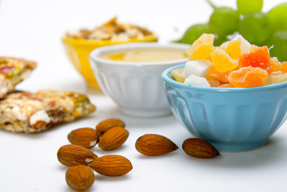 A blue bowl filled with colorful dried fruit sits next to a small white bowl of dip, surrounded by almonds and a yellow bowl of mixed nuts, with green grapes in the background on a white surface.