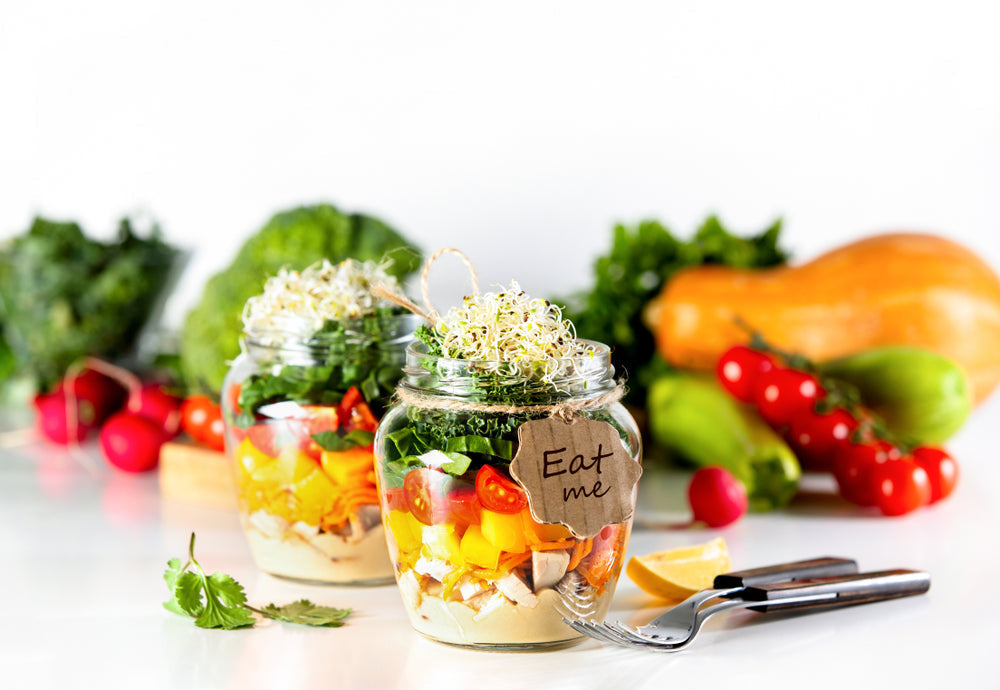 Two glass jars filled with colorful layers of vegetables and greens, topped with sprouts, sit on a white surface, accompanied by fresh produce and a tag reading "Eat me."