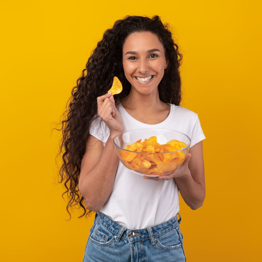 A woman with long, curly hair smiles while holding a chip in one hand and a bowl of chips in the other, set against a bright yellow background.