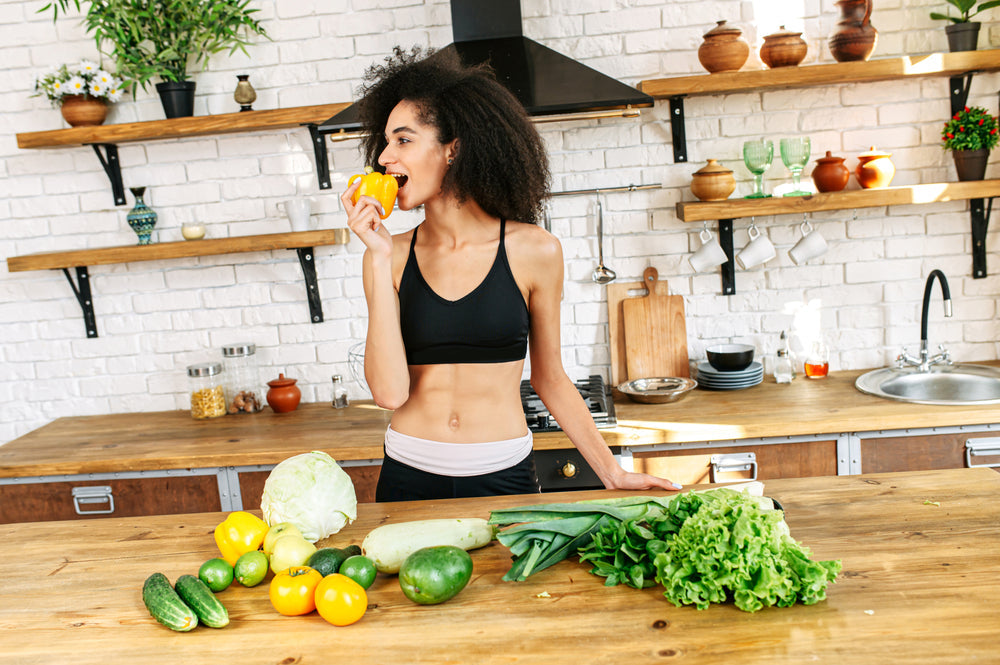 A woman in a black sports bra smiles while holding a yellow bell pepper, surrounded by various fresh vegetables on a wooden kitchen counter with shelves of dishes and plants in the background.