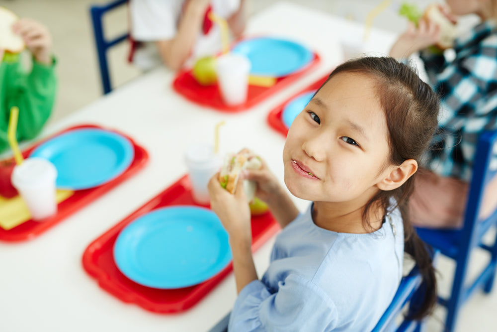 A young girl with dark hair smiles while holding a sandwich in a school cafeteria, surrounded by classmates eating lunch at tables with blue plates and drinks.
