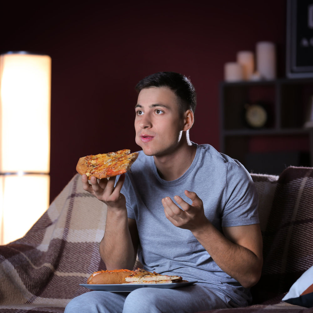 A young man in a gray shirt sits on a couch, holding a slice of pizza and looking thoughtfully at it, with a plate of pizza beside him in a cozy, dimly lit room.