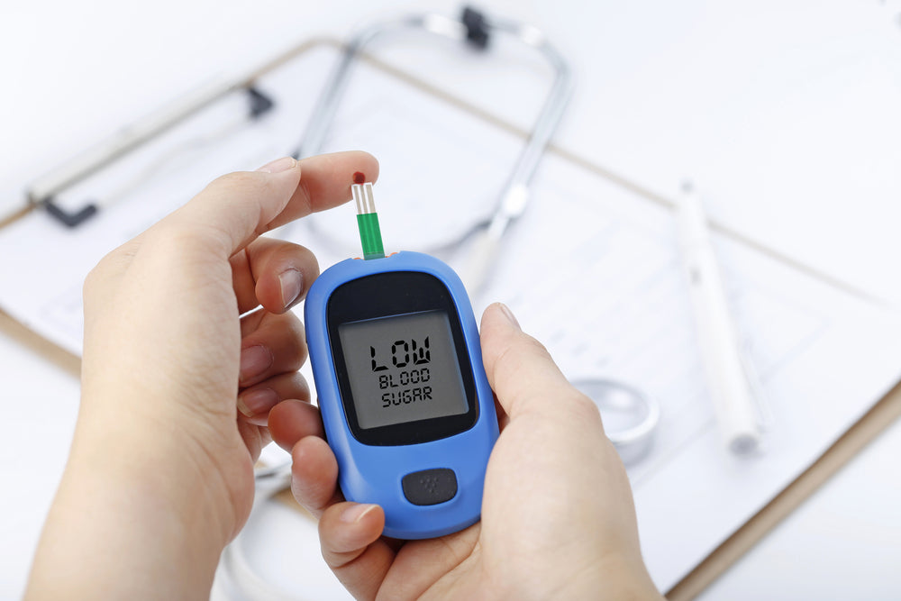 A person holds a blue blood glucose meter displaying "LOW BLOOD SUGAR," with a blood sample on the testing strip, surrounded by medical tools and paperwork in a clinical setting.